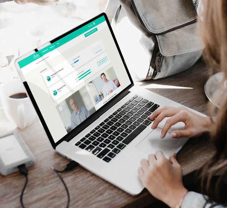 A young female sits in a coffee shop with her laptop opened and scrolling through the Better Together program displayed on her screen