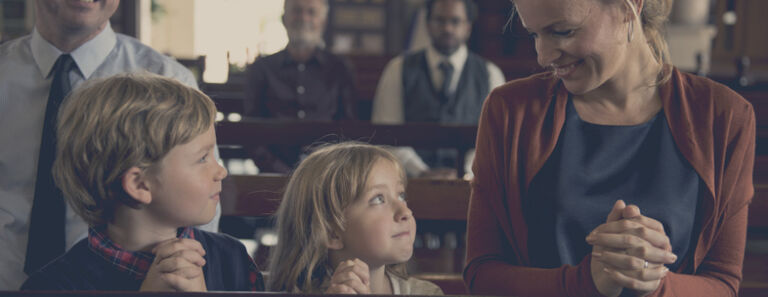 A mother with her hands crossed kneels with her daughter and son on her left side who also have their hands crossed in prayer at church 