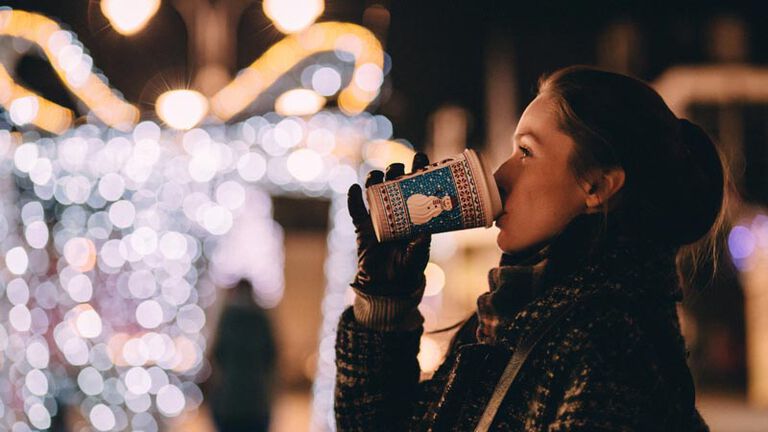 A young woman stands outside bundled up with a scarf and gloves drinking hot chocolate while looking at bright christmas lights during the advent season